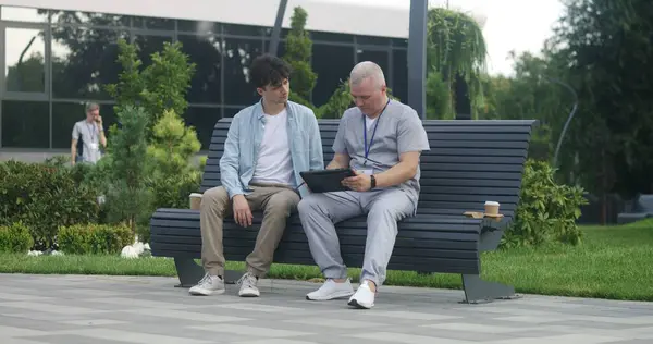 Doctor with tablet in hands sits with adult patient on bench and suggests treatment to him. Professional in medical uniform makes diagnosis to client using digital tablet. Medical staff work outdoor.