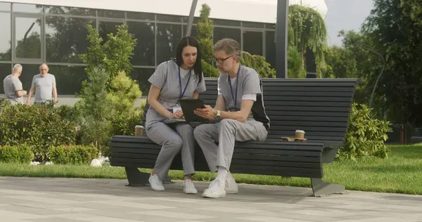 Two doctors sit on the bench and discuss patient treatment. Healthcare specialists look at client tests results using tablet. Woman helps colleague with making diagnosis. Medical staff work outdoor.