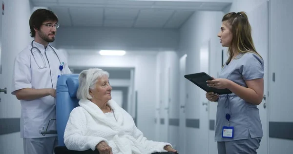 女性医師は 車椅子の医師や高齢女性と診療所の廊下に立って 検査結果を議論し デジタルタブレットを使用しています 病院の廊下でバックグラウンドを歩く医療スタッフと患者 — ストック写真