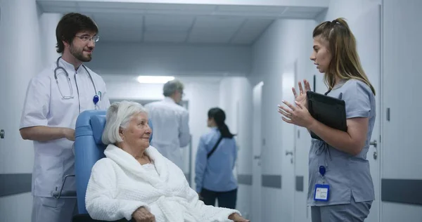 女医生与坐在轮椅上的医生和老年妇女站在诊所走廊上 讨论测试结果 使用数字平板电脑 医护人员和病人在医院走廊的背景下行走 — 图库照片