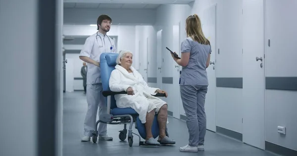 男性看護師は 診療回廊で女性患者と車椅子をプッシュします 女性医師は同僚や高齢の患者に会い 話し合い デジタルタブレットを使用する 病院の廊下の医療スタッフと患者 — ストック写真