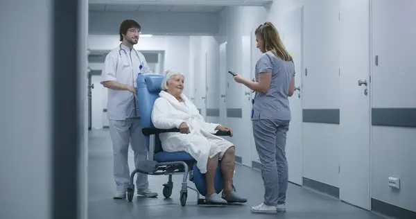 男性看護師は 診療回廊で女性患者と車椅子をプッシュします 女性医師は同僚や高齢の患者に会い 話し合い デジタルタブレットを使用する 病院の廊下の医療スタッフと患者 — ストック写真