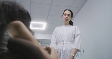 Kadın doktor klinik koridorunda duruyor, tekerlekli sandalyedeki hastaya danışıyor. Sağlık uzmanı fiziksel engelli bir kadınla tıbbi muayene hakkında konuşur. Modern tıp merkezinin koridoru..