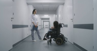 Kadın doktor klinik koridorunda duruyor. Tekerlekli sandalyede omurilik kası atrofisi olan bir kadına danışıyor. Doktor, engelli bir hastayla sağlık kontrolü hakkında konuşur. Modern tıp merkezinin koridoru..