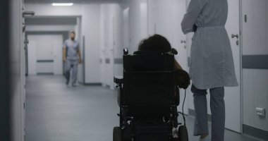 Kadın doktor klinik koridorunda yürüyor, fiziksel engelli bir kadına danışıyor. Doktorlar elektrikli sandalyedeki hastalara tıbbi prosedürlerden bahseder. Modern tıp merkezinin koridoru..