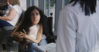 Elektrikli sandalyedeki kadın hastane ya da klinik lobisinde doktorla konuşuyor. Kadın hekim, fiziksel engelli bir hastaya danışıyor. Modern tıp merkezinin bekleme salonunda insanlar kanepeye oturur..