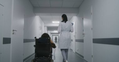 Kadın doktor klinik koridorunda yürüyor, fiziksel engelli bir kadına danışıyor. Doktorlar elektrikli sandalyedeki hastalara tıbbi prosedürlerden bahseder. Modern tıp merkezinin koridoru..