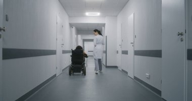 Kadın doktor klinik koridorunda yürüyor, fiziksel engelli bir kadına danışıyor. Doktorlar elektrikli sandalyedeki hastalara tıbbi prosedürlerden bahseder. Modern tıp merkezinin koridoru..