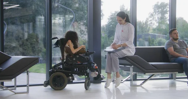 Woman in wheelchair talks to doctor in hospital or clinic lobby. Female physician sits on couch, consults patient with physical disability, uses mobile phone. Waiting area of modern medical facility.