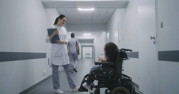 女性医師は診療所の廊下に立って 車椅子で脊髄筋萎縮の女性に相談する 医師は 医療検査について障害のある患者と話す 現代医療センターのホールウェイ — ストック写真