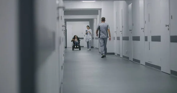 女医生走在诊所走廊上 为身体残疾的病人提供咨询 医生在谈论与坐汽车轮椅的妇女进行体检 现代医疗设施的走廊 — 图库照片