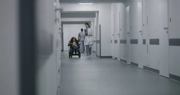 Female doctor walks down the clinic corridor, consults patient with physical disability. Physician talks about medical checkup with woman in motorized wheelchair. Hallway of modern medical facility.