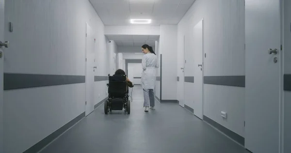 Female doctor walks down the clinic hallway, consults woman with physical disability. Physician talks about medical procedures with patient in electric wheelchair. Corridor of modern medical facility.