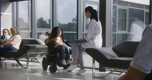 坐在机动轮椅上的残疾妇女在现代医院或诊所大堂与医生交谈 专业医生坐在沙发上 为脊柱肌肉萎缩的女性患者提供咨询 Dolly Shot — 图库照片