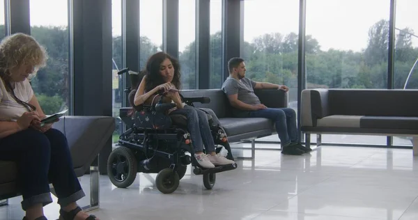 Woman with SMA disability in electric wheelchair waits for consultation with doctor in clinic waiting area, uses mobile phone. Diverse patients sit on sofas in hospital or modern medical center lobby.