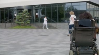 Fiziksel engelli bir kadın doktorla randevusu olan motorlu tekerlekli sandalyeyle kliniğe gidiyor. Hastane personeli ve çeşitli kişiler hastaneye girip çıkıyor. Modern tıp merkezinin avlusu..