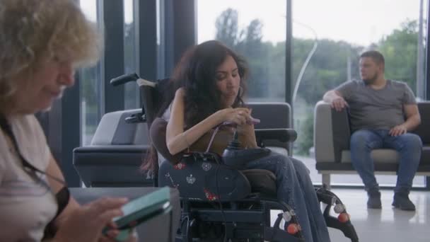 坐轮椅的残疾妇女在诊所大厅等医生的预约 使用手机 不同的人坐在医院或现代医疗设施等候区的沙发上 — 图库视频影像
