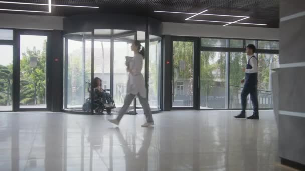 患有Sma残疾的妇女乘坐机动轮椅通过旋转门进入诊所入口 男管理员与女病人见面并打招呼 医院或现代医疗设施大堂 — 图库视频影像