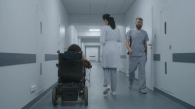 Kadın doktor klinik koridorunda yürüyor, engelli bir kadına danışıyor. Doktorlar tekerlekli sandalyedeki hastalara tıbbi prosedürlerden bahseder. Tıbbi tesis koridoru. İzleme görüntüsü. Arka plan.