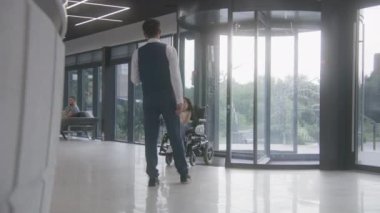 Fiziksel engelli bir kadın, resepsiyona elektrikli tekerlekli sandalyeyle gidiyor. Sağlık çalışanı hastaları karşılar ve selamlar. Modern tıp merkezinin bekleme salonunda çeşitli insanlar koltuklarda oturur..