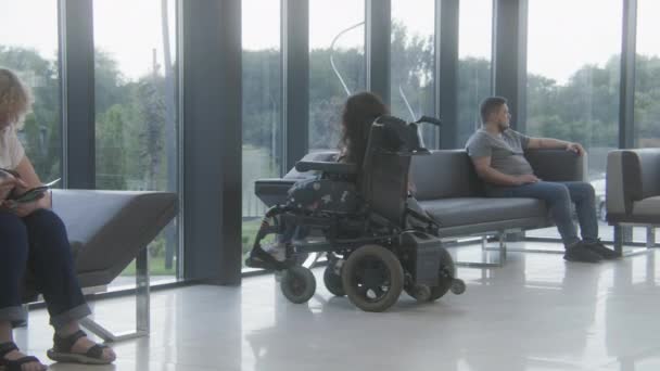 身体残疾的妇女乘坐机动轮椅在诊所大堂 等待医生 使用手机 不同的人坐在医院或现代医疗中心候诊室的沙发上 — 图库视频影像