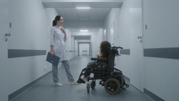 女医生站在诊所走廊里 为轮椅上的脊髓肌肉萎缩的妇女提供咨询 医生与残疾病人谈论体格检查 现代医疗中心的走廊 — 图库视频影像