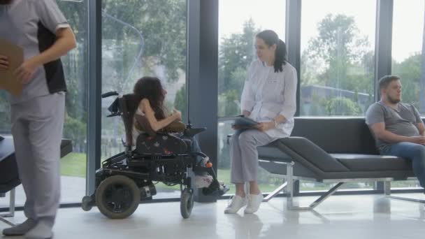 坐轮椅的妇女在医院或诊所大堂与医生交谈 女医生坐在沙发上 为身体残疾的病人提供咨询 使用手机 现代医疗设施候诊区 — 图库视频影像