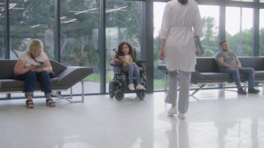 Elektrikli sandalyedeki kadın klinik bekleme salonunda doktorla görüşmek için bekliyor. Hemşire, engelli hastaları ameliyatlara davet ediyor. Tıbbi personel ve modern tıp merkezinin lobisindeki insanlar..