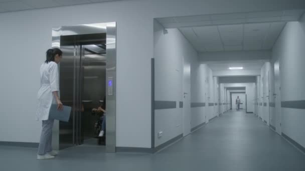 坐着机动轮椅的妇女和医生一起离开电梯 沿着诊所走廊骑马 女医生会诊患有Sma残疾的病人 在现代医院或医疗设施的走廊上行走 — 图库视频影像