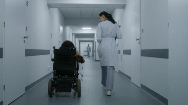女医生走在诊所走廊上 为身体残疾的妇女提供咨询 医生说要给坐轮椅的病人做身体检查 现代医疗中心的走廊 慢动作 — 图库视频影像