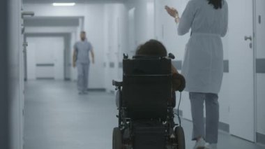 Kadın doktor klinik koridorunda yürüyor, fiziksel engelli bir kadına danışıyor. Doktorlar elektrikli sandalyedeki hastalara tıbbi prosedürlerden bahseder. Modern tıp merkezinin koridoru..