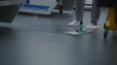 Yetişkin hemşire yeni hastalar için hastane koğuşunu temizliyor, yerleri paspaslıyor. Sağlık çalışanı temizlik arabasına süpürge koyar ve halı alır. Hastalardan sonra temizlikçi hastane odasını temizler. Klinikte çalışan tıbbi personel.