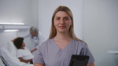 Yetişkin hemşire elinde dijital tablet bilgisayarla hastane odasında kameraya bakıyor. Olgun kadın doktor arka plandaki hastanın durumunu kontrol ediyor. Modern tıp merkezinde tıbbi personel.