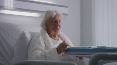 Yaşlı kadın lezzetli yemekler yiyor ve parlak hastane koğuşundaki yatakta dinlenen kameraya bakıyor. Başarılı bir ameliyattan sonra iyileşen yaşlı kadın hasta. Modern tıbbi tesis ya da klinik. Portre.