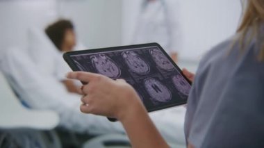 Kadın doktor hastane odasında duruyor, MR veya CT tarama sonuçlarını incelemek için dijital tablet bilgisayar kullanıyor. Ergen yatakta yatıyor ve arka planda doktorla konuşuyor..