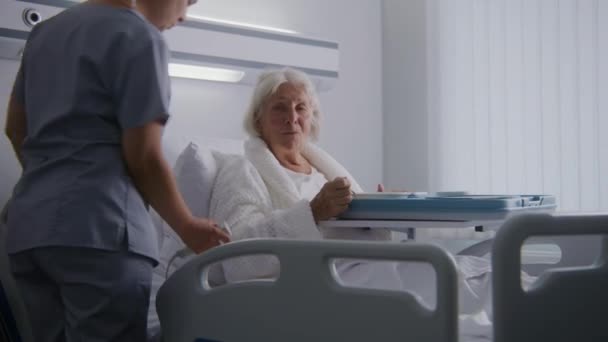 老年妇女在明亮的病房里吃着美味的食物 手术成功后正在康复 友好的护士为老年女性病人铺床 现代医疗设施或诊所 — 图库视频影像