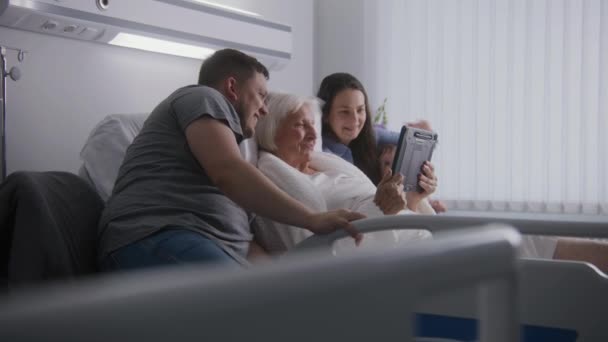 病院の部屋 愛する家族は 手術が成功した後に回復する高齢のおばあちゃんをサポートします デジタルタブレットを使用したビデオ通話による親戚の話 一緒に時間を過ごす 現代医療施設 クリニック — ストック動画
