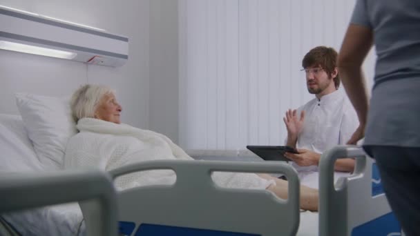 医生会诊在病房卧床休息的老年女性病人 男医生使用数码平板电脑 与护士交谈 老年妇女在手术成功后康复 现代诊所 — 图库视频影像