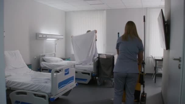 卫生工作者把清洁推车送到医院病房 护士在病人之后清洁医院房间 和她同事一起打扫房间的人在床上换床 在现代诊所工作的医务人员 — 图库视频影像