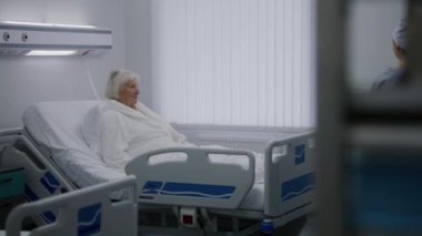 Üniformalı hemşire parlak hastane odasına giriyor, yaşlı kadın hastalara yemek ve yemek getiriyor. Başarılı bir ameliyattan sonra yaşlı kadın yatakta dinleniyor. Modern tıbbi tesis veya klinik.