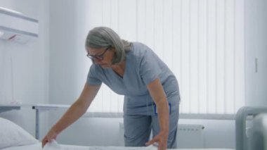 Profesyonel kadın temizlikçi hastane odasında yatak örtülerini değiştiriyor. Olgun hemşire hastane koğuşunda yatağı hazırlar ve yeni hastalar için hazırlar. Modern tıp merkezinde çalışan tıbbi personel..