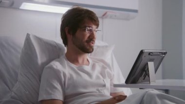 Yetişkin bir adam hastane odasında yatar ve ortaklarıyla online konferansta tablet kullanarak konuşur. Gözlüklü adam uzaktan çalışıyor. Hasta başarılı bir ameliyattan sonra tamamen iyileşiyor. Modern parlak klinik.