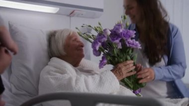 Küçük çocuklu bir aile başarılı bir ameliyattan sonra iyileşen büyükannesini ziyaret eder ve çiçek verir. Yaşlı kadın, parlak hastane koğuşunda yatıp dinleniyor. Modern tıbbi tesis veya klinik.