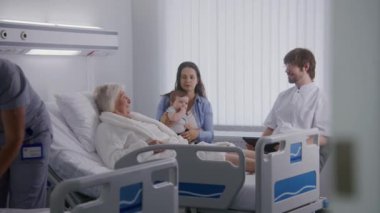 Yaşlı kadın ve ailesi hastane koğuşunda. Erkek hemşire, doktor tablet bilgisayar kullanıyor, kadın kıdemli hastaya danışmanlık yapıyor ve başarılı bir ameliyattan sonra iyileşiyor. Modern tıbbi tesis.
