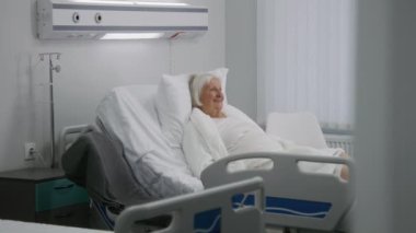 Yaşlı kadın hasta, aydınlık hastane odasında yatağında dinleniyor. Aile ziyaretleri başarılı bir ameliyattan sonra iyileşen büyük anne, çiçek verir. Modern tıbbi tesis ya da klinik. Yavaş çekim.