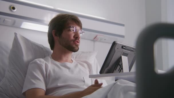 成年男子躺在医院病房的床上 用平板电脑在网上会议上向客户咨询 戴眼镜的人远程工作 手术成功后 病人完全康复 现代明亮的诊所 — 图库视频影像