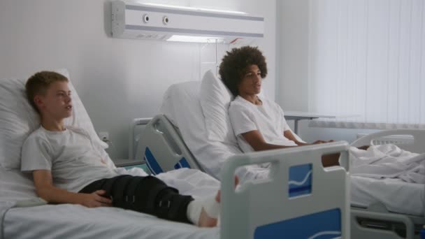 非裔美国青少年与室友讨论电影 带绷带腿的小男孩躺在医院房间的床上 手术成功后完全康复的病人 现代明亮的医疗中心 — 图库视频影像