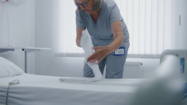 Parlak Hastane Koğuşunda Kadın Temizlikçi Yatak Örtülerini Değiştiriyor Olgun Hemşire — Stok video