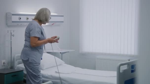 成熟的护士在医院病房里用遥控装置为病人调整病床 女医务工作者推着轮椅和老妇人在一起 把她放在床上 现代诊所或医疗设施的医务人员 — 图库视频影像