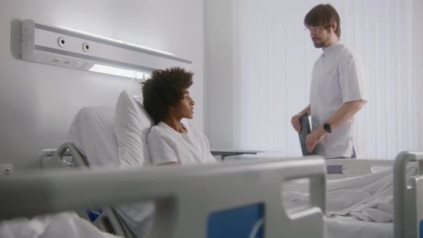 躺在床上的小男孩的护士绷带腿 手握平板电脑的医生与非洲裔美国青少年讨论手术结果 手术成功后病人康复 明亮的病房 — 图库视频影像